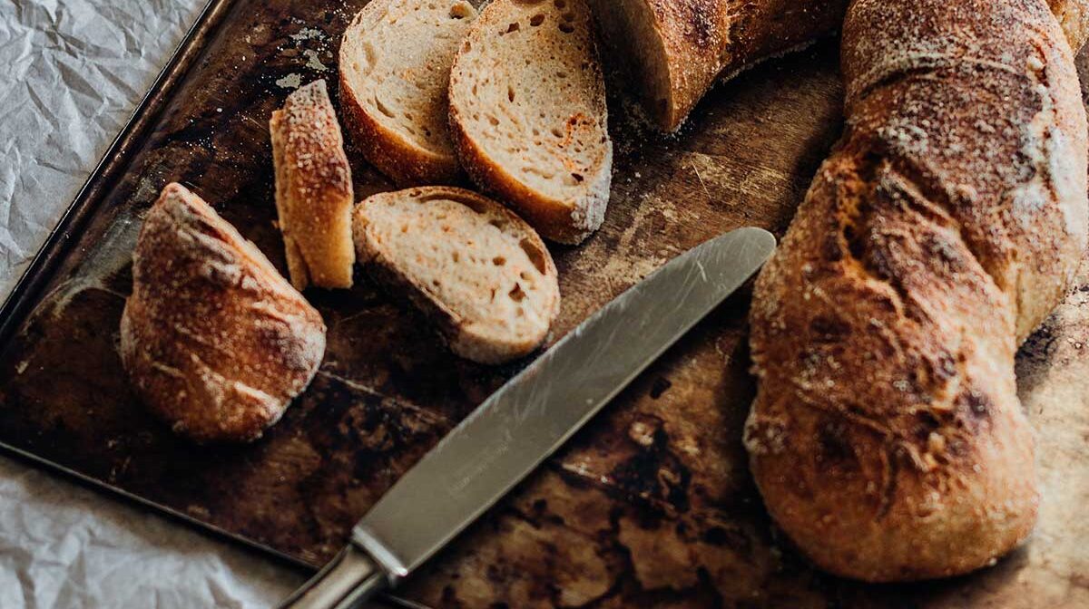 h4-blog6 Sourdough baguette sliced and displayed on a weathered metal tray for a minimalist bakery catalog