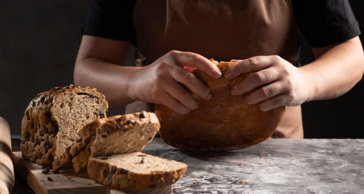 h4-blog7 Baker's hands breaking fresh sourdough bread next to sliced multi-seed loaf on a rustic stone counter