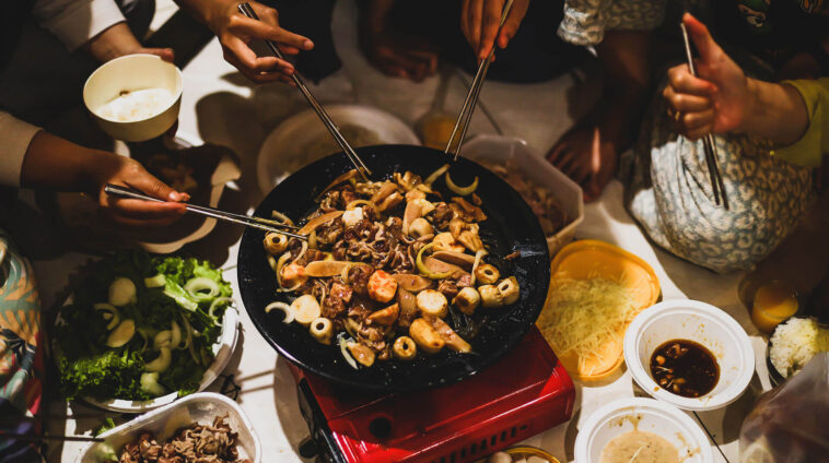 A group of friends using chopsticks to cook Korean BBQ or stir-fry on a portable red gas stove at a social gathering.