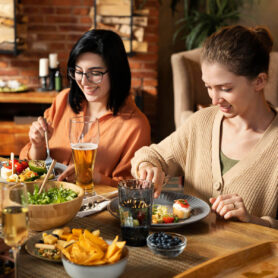 Two young women smiling and eating a healthy dinner with friends, featuring salad, appetizers, wine, and beer in a rustic dining room.