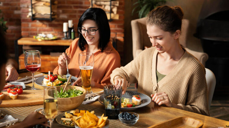 Two young women smiling and eating a healthy dinner with friends, featuring salad, appetizers, wine, and beer in a rustic dining room.