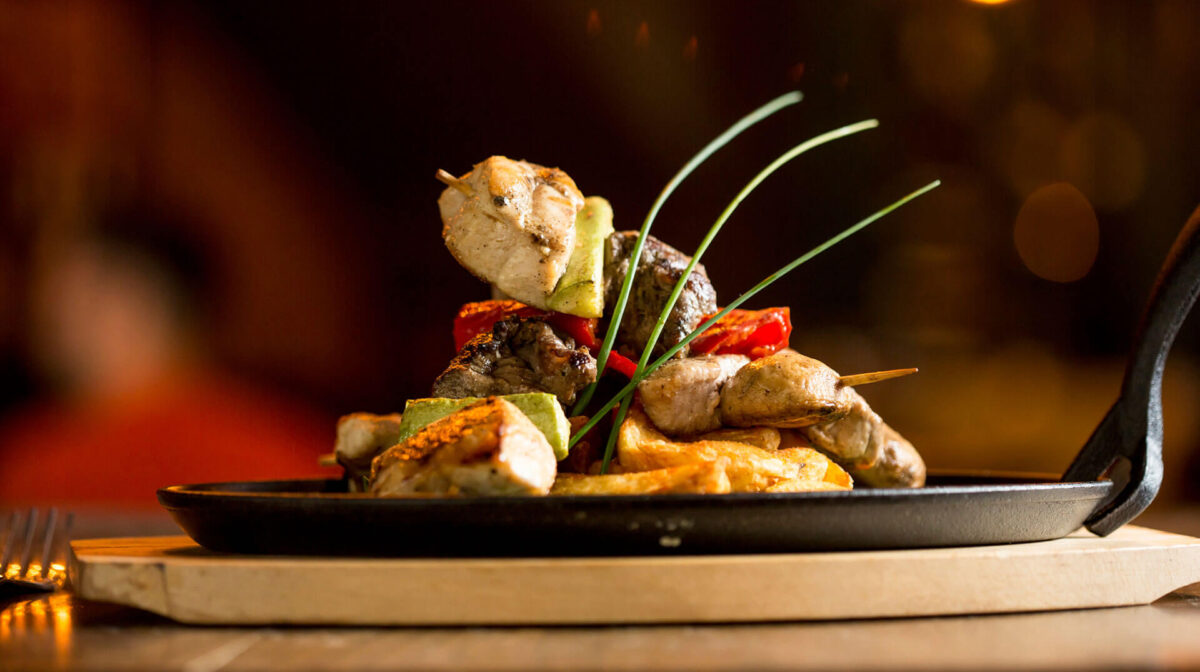 Mediterranean-style grilled chicken and beef skewers with roasted potatoes and fresh herbs in a restaurant setting.
