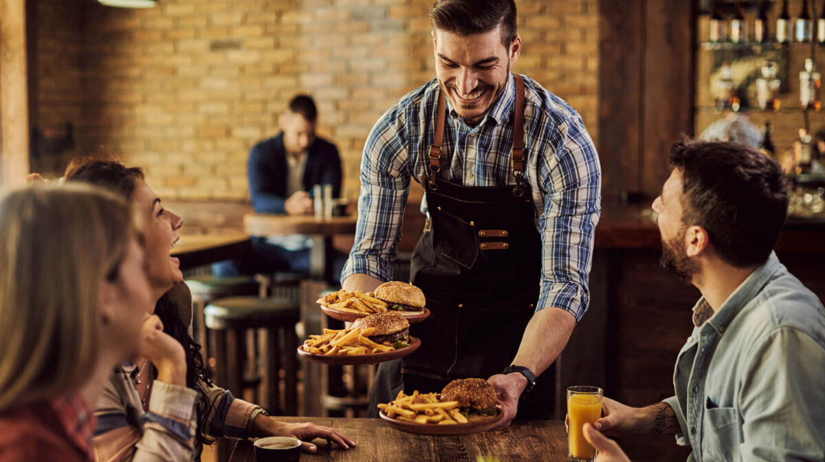 theme-05 A smiling waiter in a plaid shirt and apron serving three plates of burgers and fries to a group of friends at a rustic restaurant table.