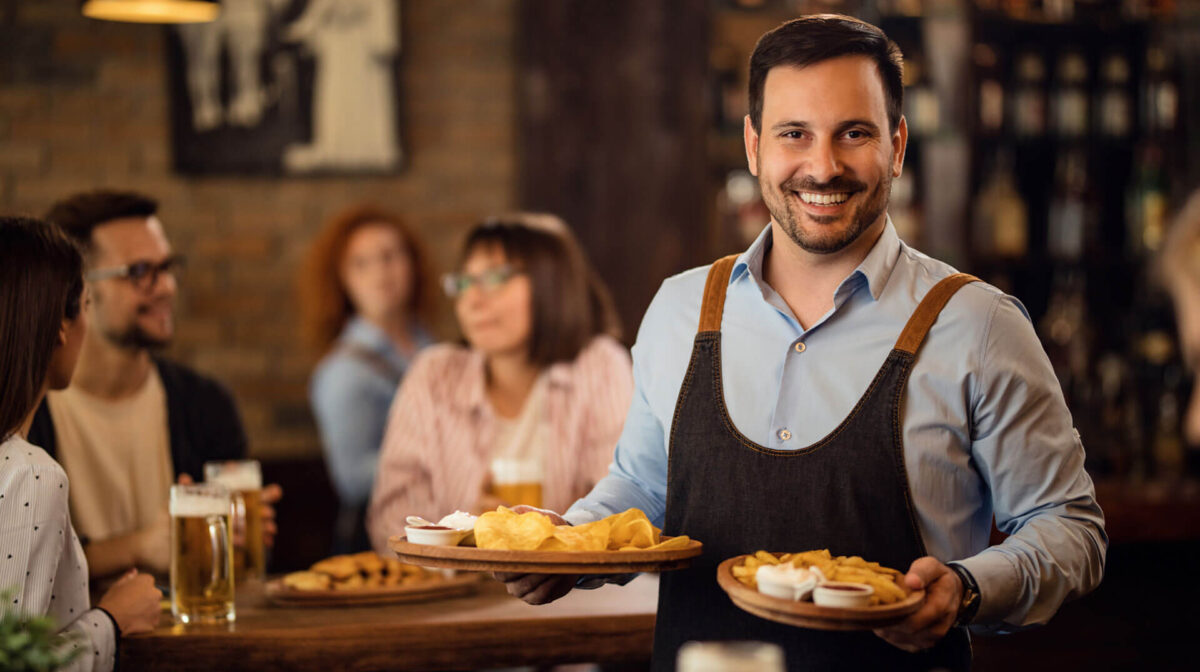 theme-06 A smiling male waiter in a blue shirt and denim apron serving trays of chips and appetizers to customers in a cozy pub.
