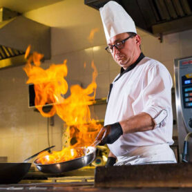 Professional chef in a white uniform sautéing vegetables with a large flame in a commercial kitchen