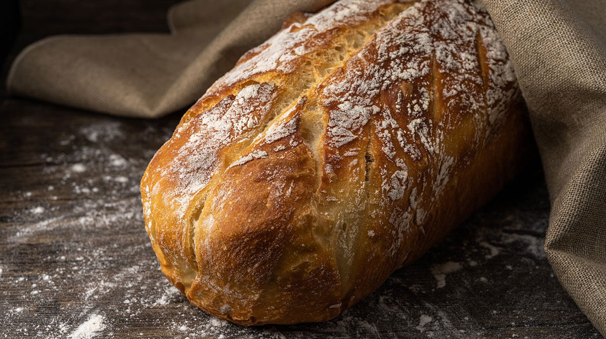h4-blog1 Rustic sourdough bread loaf with a flour-dusted crust resting on a weathered wooden table with burlap fabric