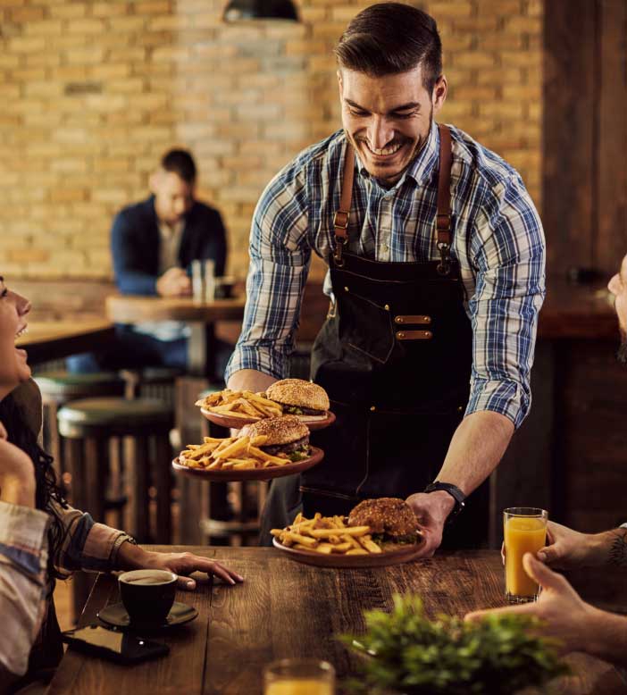 Smiling waiter serving gourmet burgers and fries to customers in a modern restaurant with rustic brick decor